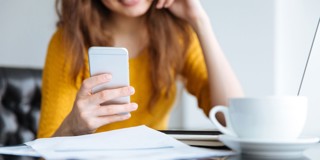 Vrouw in gele trui kijkt op haar smartphone terwijl ze aan een tafel zit met papieren en een kopje koffie.