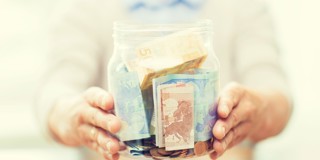 Handen houden een glazen pot vast met een mix van munten en biljetten.