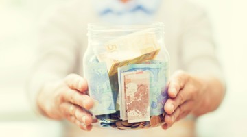 Handen houden een glazen pot vast met een mix van munten en biljetten.