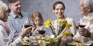 Een groep mensen zit aan tafel en geniet van een diner met salade, rode wijn en op de achtergrond een vaas met gele narcissen.