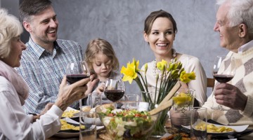 Een groep mensen zit aan tafel en geniet van een diner met salade, rode wijn en op de achtergrond een vaas met gele narcissen.