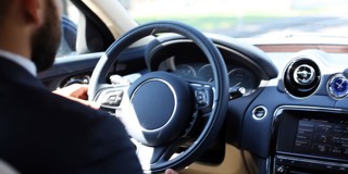Een man in een zwart pak, met zijn handen aan het stuur van een luxe auto, kijkt gefocust voor zich uit.