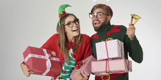 Twee mensen in feestelijke kleding houden cadeaus vast, omringd door een bel en kerstslingers in een feestelijke sfeer.