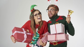 Twee mensen in feestelijke kleding houden cadeaus vast, omringd door een bel en kerstslingers in een feestelijke sfeer.