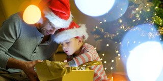 Een volwassene en een kind met kerstmanmutsen kijken naar een cadeau dat met gouden inpakpapier is verpakt, omringd door feestelijke verlichting.