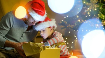 Een volwassene en een kind met kerstmanmutsen kijken naar een cadeau dat met gouden inpakpapier is verpakt, omringd door feestelijke verlichting.
