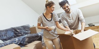 Een vrouw en een man openen verhuisdozen in een lege woonkamer vol inpakdozen. De vrouw heeft een kom in haar hand.