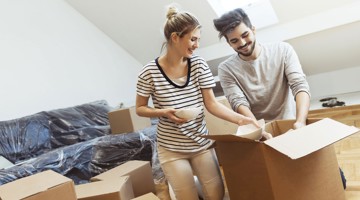 Een vrouw en een man openen verhuisdozen in een lege woonkamer vol inpakdozen. De vrouw heeft een kom in haar hand.