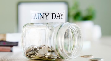 Glazen pot met munten en een label met de tekst RAINY DAY, als symbool voor een spaarpot voor noodgevallen.