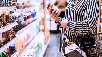 Vrouw in gestreepte blouse houdt een pot pastasaus vast in de gang van een supermarkt, met een winkelmandje naast zich.