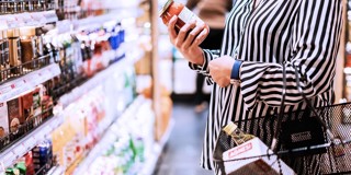 Femme en blouse rayée vérifiant un pot de sauce tomate tout en tenant un chariot de supermarché.