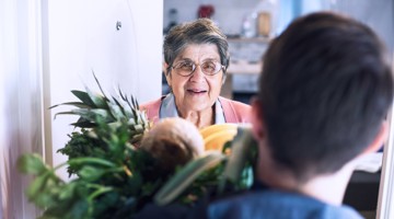 Une personne se tient devant la porte avec un sac de courses rempli de légumes et de fruits, tandis qu'une autre personne ouvre la porte.