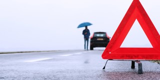 Une personne avec un parapluie se tient près d'une voiture en panne, à côté d'un triangle de signalisation rouge sur une route mouillée.