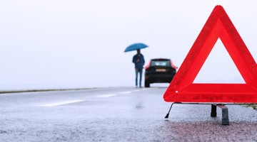 Une personne avec un parapluie se tient près d'une voiture en panne, à côté d'un triangle de signalisation rouge sur une route mouillée.
