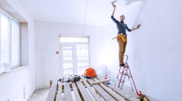 Man op een ladder die een muur schildert in een onafgebouwde ruimte, met gereedschap en een veiligheidshelm op de vloer.