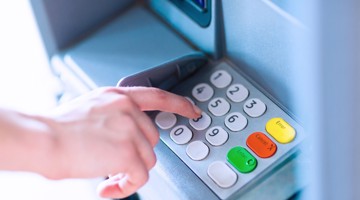 Une main appuie sur le clavier d'un distributeur automatique de billets, avec des chiffres et des fonctions, y compris un bouton vert et un bouton jaune.