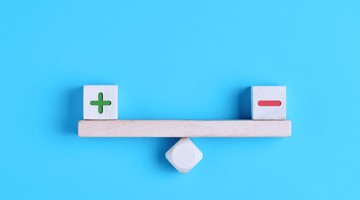 Houten balans met een groen plusteken en een rood minteken aan beide zijden, tegen een blauwe achtergrond.
