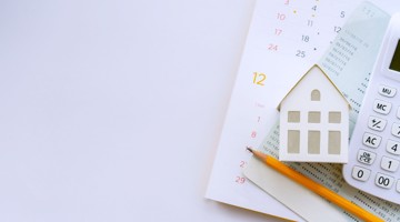 Huisvormige figuur op een kalender, vergezeld van een rekenmachine en een potlood, voor financiële planning of vastgoedbeheer.