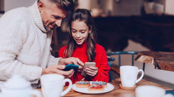 Un adulte et un enfant sont assis à une table avec des pâtisseries et deux tasses de café, tous deux regardant leurs téléphones.