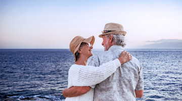 Un couple portant des chapeaux s'étreint sur la côte, avec l'océan en arrière-plan et un ciel coloré au coucher du soleil.