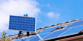 Technicus installeert een zonnepaneel op een dak, omringd door andere zonnepanelen onder een heldere blauwe lucht.