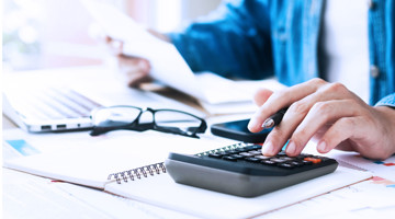 Homme tenant un stylo, utilisant une calculatrice tout en consultant des notes sur un bureau avec un ordinateur portable et un carnet.