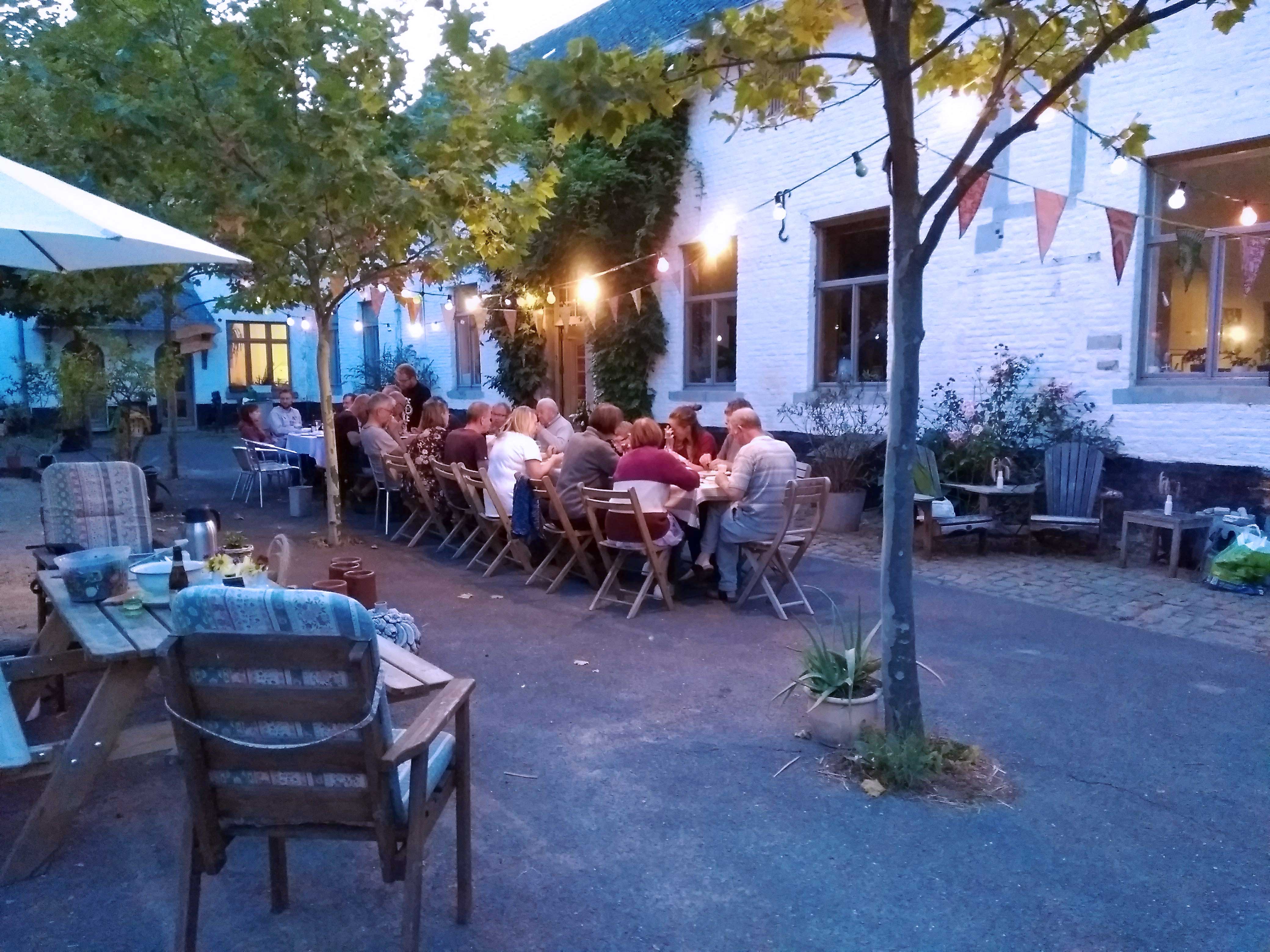 Een lange tafel met mensen die samen dineren in een sfeervolle buitenruimte, omringd door bomen en feestelijke verlichting.