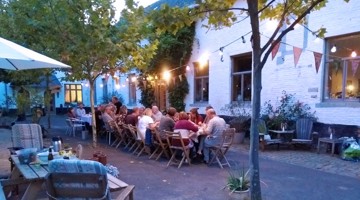 Een lange tafel met mensen die samen dineren in een sfeervolle buitenruimte, omringd door bomen en feestelijke verlichting.