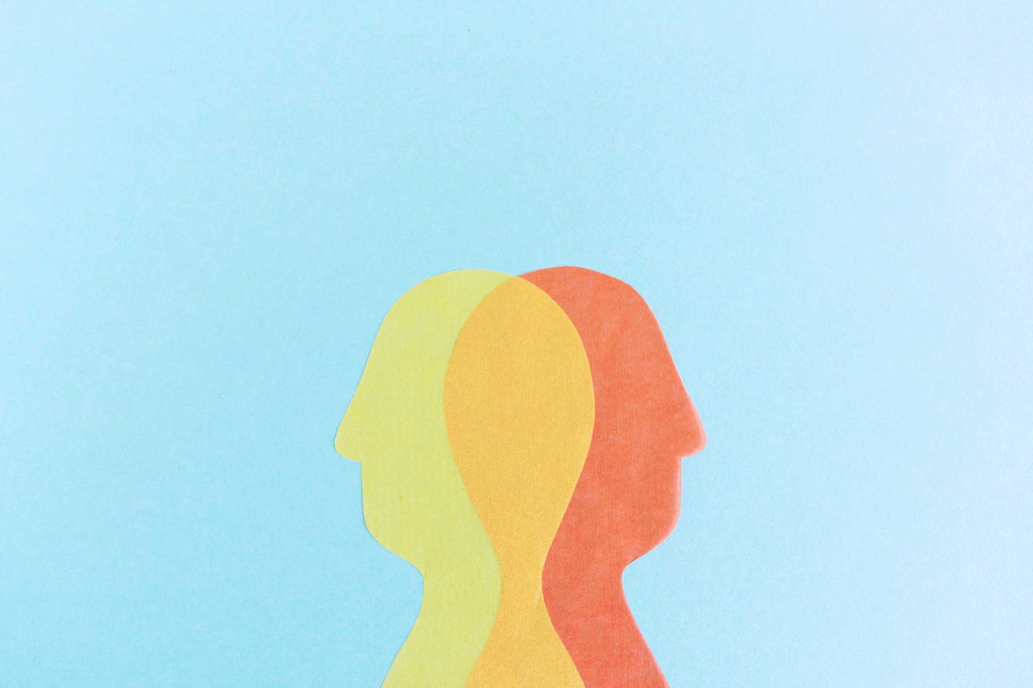 Silhouetten van twee overlappende gezichten in geel en oranje, op een lichtblauwe achtergrond, die samenwerking of communicatie symboliseren.