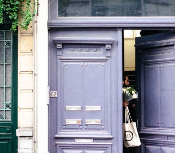 Une personne avec un bouquet de fleurs entre par une double porte violette, avec une porte verte à gauche.