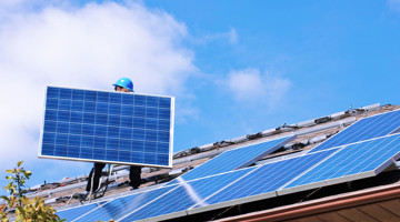 Un homme portant un casque bleu installe des panneaux solaires sur un toit, avec un ciel bleu clair en arrière-plan.