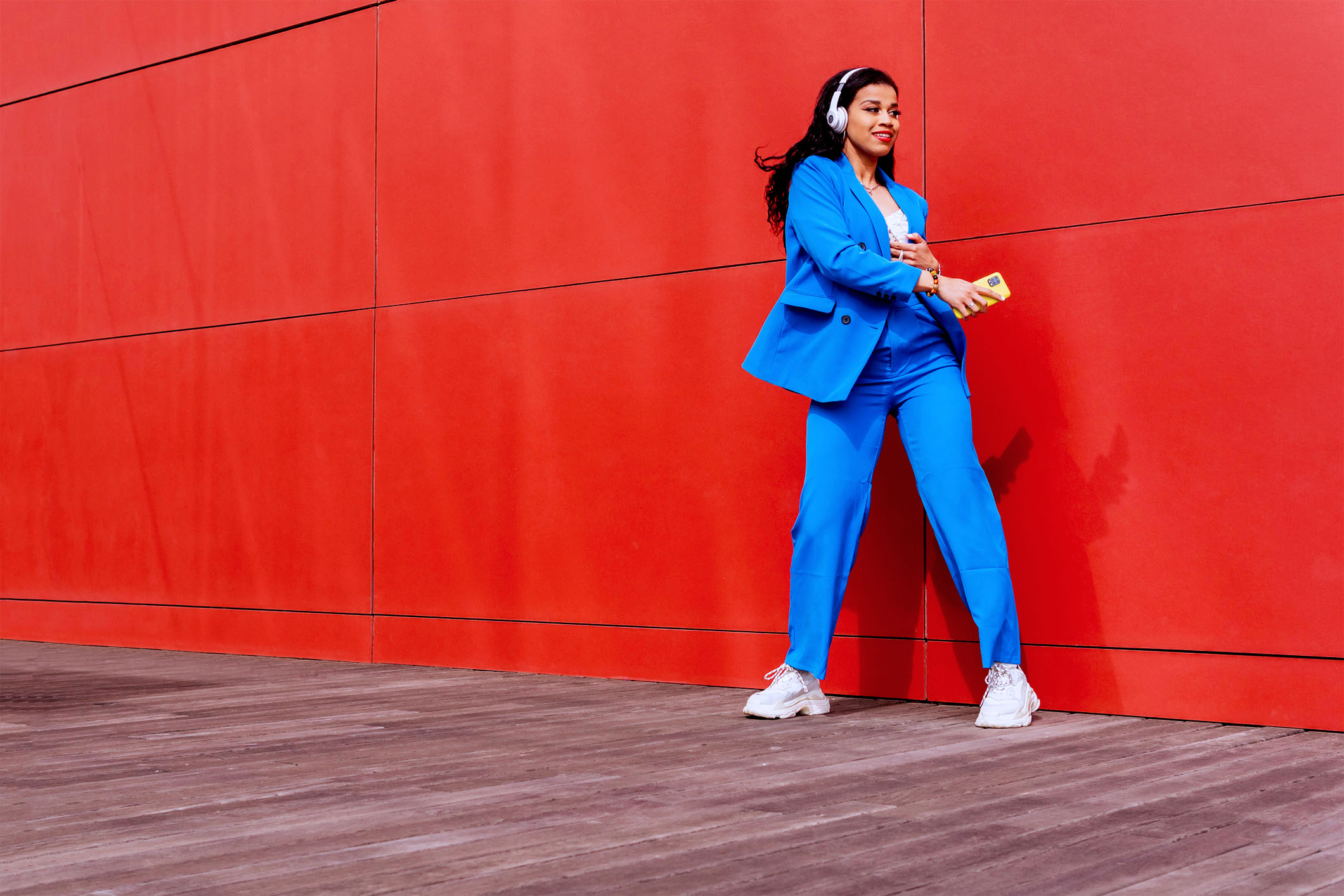 Une femme en blazer bleu et pantalon assorti se tient détendue contre un mur rouge vif, écoutant de la musique avec des écouteurs et tenant un smartphone dans sa main.