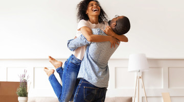 Een man tilt een vrouw op in een lichte, moderne woonkamer vol vrolijkheid.