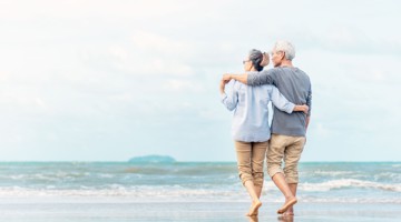 Un couple âgé se promène main dans la main sur la plage, avec la mer en arrière-plan.
