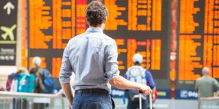 Homme avec une valise regardant un panneau d'information dans une gare, affichant les correspondances et les horaires de départ en orange.