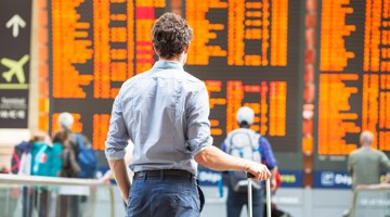 Homme avec une valise regardant un panneau d'information dans une gare, affichant les correspondances et les horaires de départ en orange.