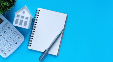 Carnet blanc avec un stylo sur fond bleu, à côté d'une calculatrice et d'une maison en papier.