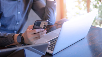 Une personne en tenue décontractée regarde son smartphone tout en étant assise à un bureau avec un ordinateur portable.