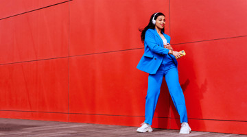 Vrouw in een blauw pak met koptelefoon leunt ontspannen tegen een rode muur en kijkt tevreden naar haar smartphone.