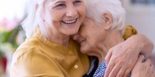 Une jeune femme étreint une femme âgée dans un cadre chaleureux avec des plantes et des fleurs en arrière-plan.
