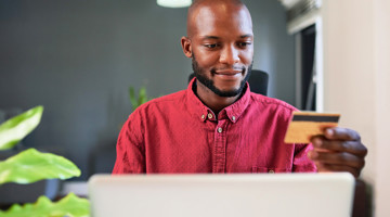 Persoon in een rode blouse gebruikt een laptop en houdt een creditcard vast, waarschijnlijk om online te winkelen.