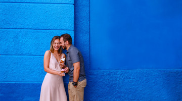 Een stel staat dicht bij elkaar tegen een heldere blauwe muur, terwijl de vrouw een ijsje vasthoudt.