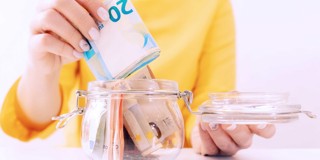 Hand plaatst een stapel van 20 euro biljetten in een glazen spaarpot, met meer bankbiljetten zichtbaar in de pot.