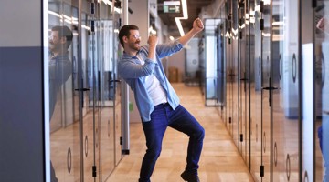 Un homme danse joyeusement dans un hall de bureau avec des murs en verre et un sol en bois.