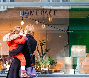 Moeder met een kind in een rode jas loopt voorbij een meubelwinkel met 'HOMEPAGE' op de etalage.