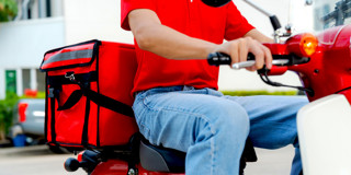 Bezorger op een rode scooter met een grote rode koeriertas, rijdt door een stedelijke omgeving.