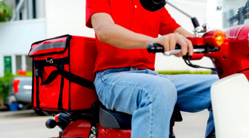 Bezorger op een rode scooter met een grote rode koeriertas, rijdt door een stedelijke omgeving.