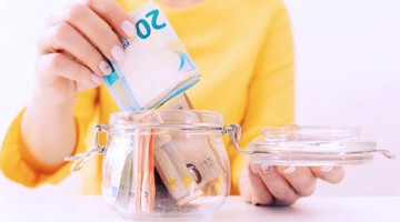 Main plaçant une pile de billets de 20 euros dans une tirelire en verre déjà remplie d'argent.