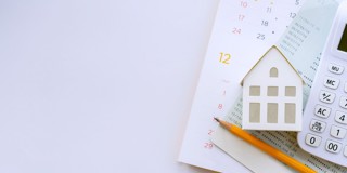 Une maison en papier repose sur un calendrier, à côté d'une calculatrice et d'un crayon.