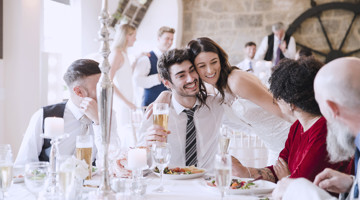 Een feestelijke tafel met gasten die met champagne proosten, omringd door een gezellige sfeer en een bruiloftssetting op de achtergrond.